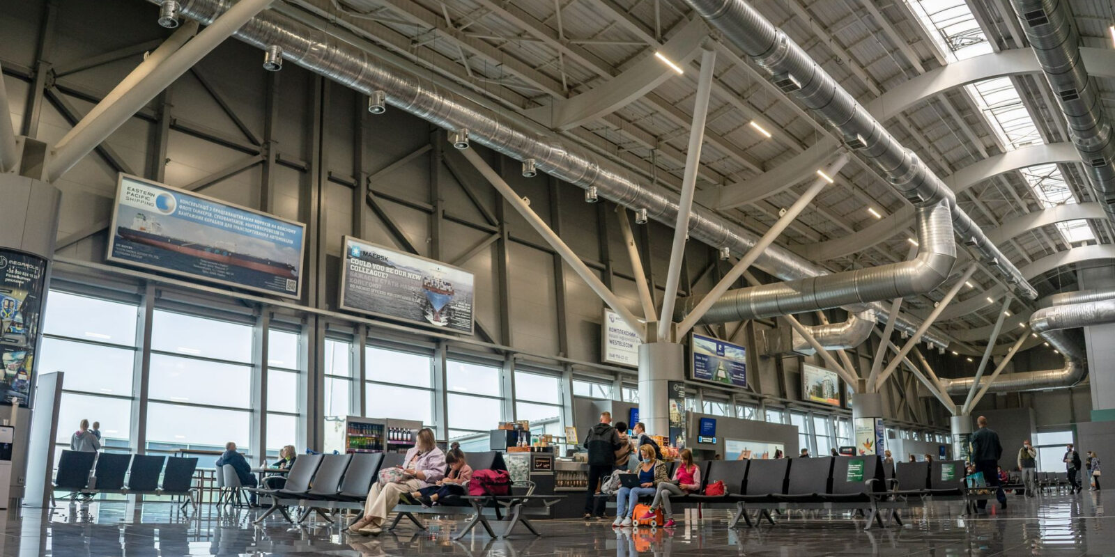 Spacious airport terminal interior with passengers seated and walking, showcasing modern design and large windows.