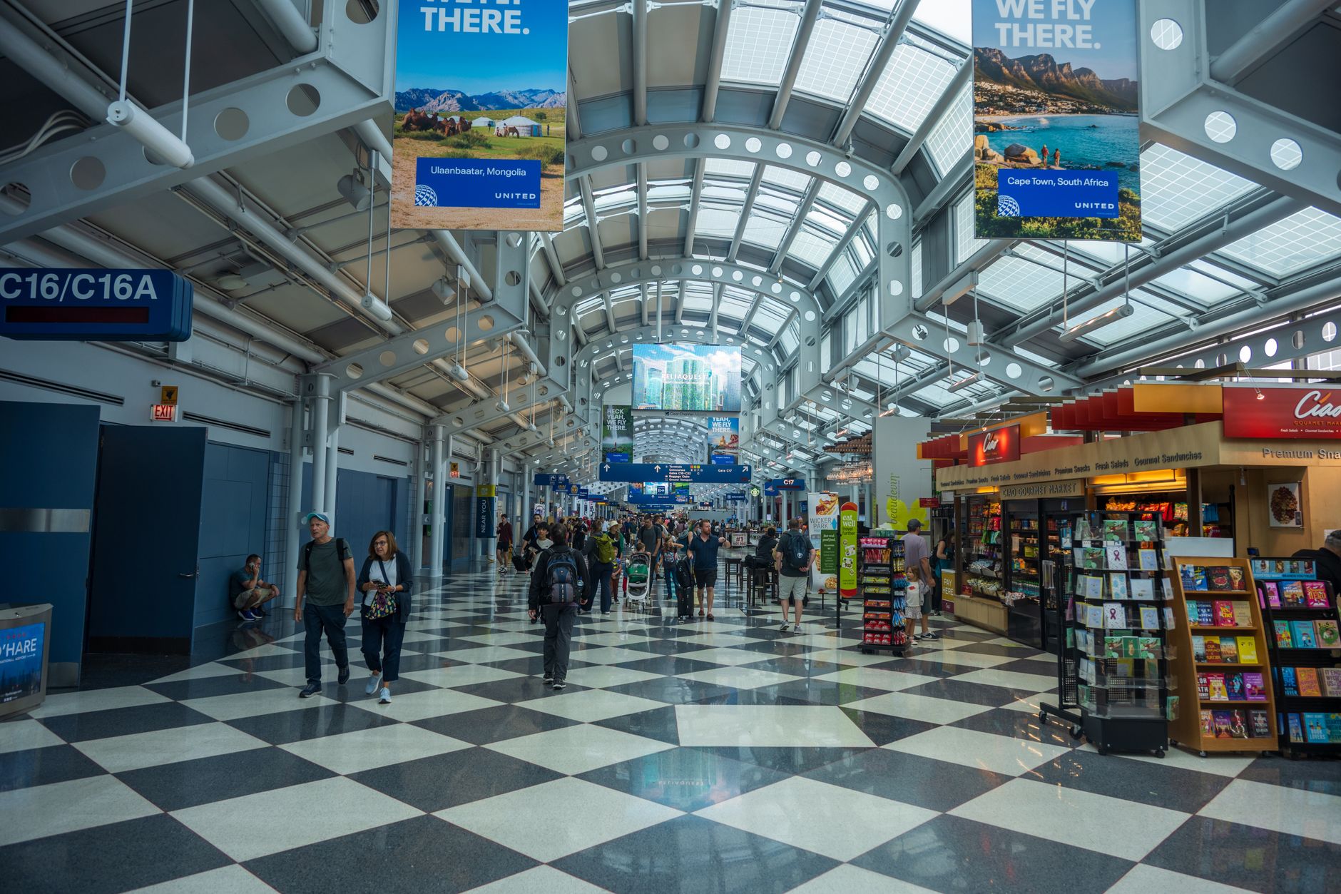 Interior view of a bustling airport terminal with travelers, shops, and overhead advertising banners.